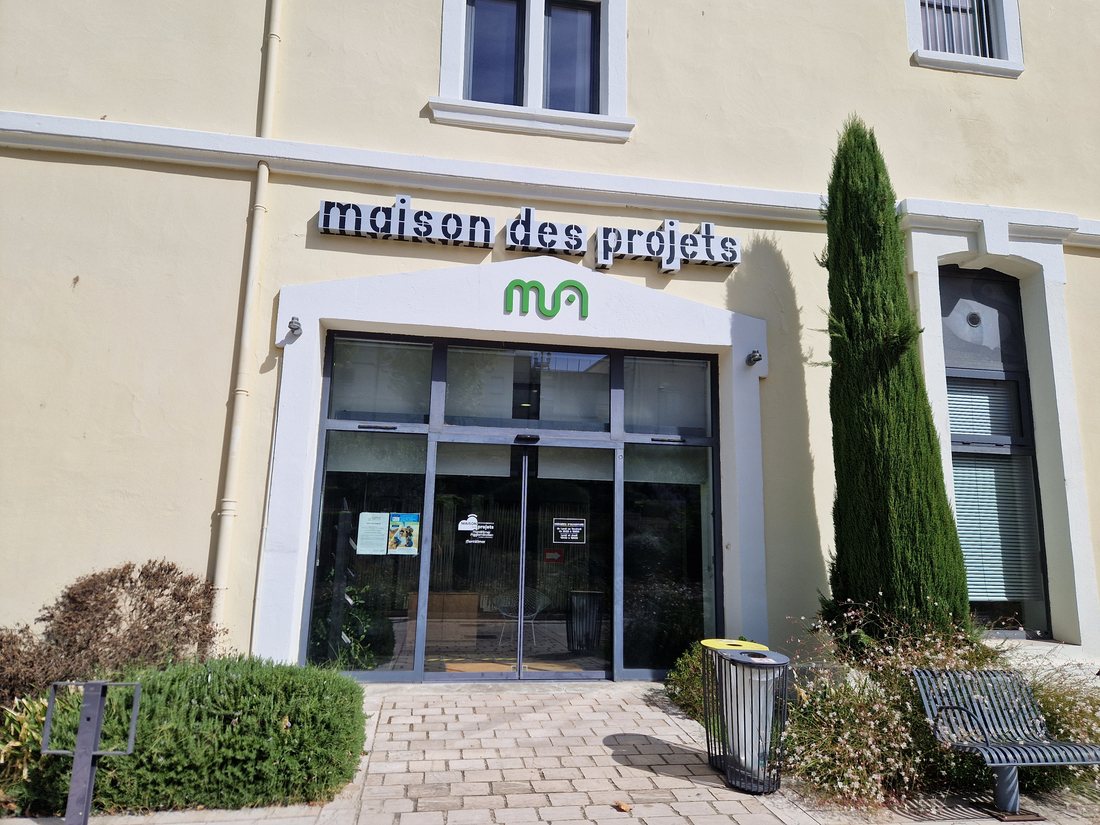 L'entrée de la Maison des projets de Montélimar Agglomération, porte vitrée sous les lettres découpées maison des projets et le logo vert de l'agglomération, avec un cyprès et une poubelle de tri