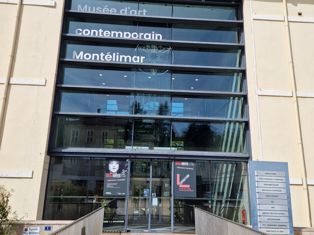 L'entrée vitrée du musée d'art contemporain de Montélimar, passerelle en bois menant à une façade de verre avec le nom du musée en lettres blanches et l'affiche de l'exposition Nicolas Bouvier