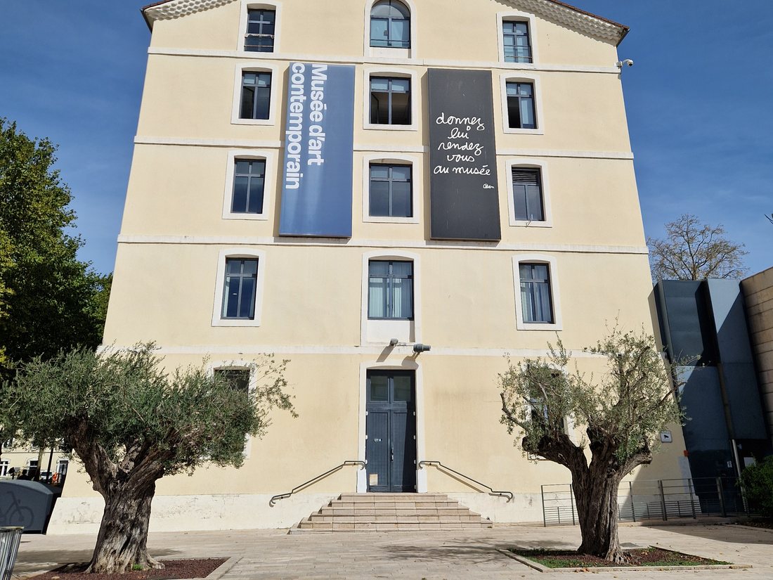 La façade du musée d'art contemporain de Montélimar, bâtiment jaune de l'ancienne caserne Saint-Martin, avec deux oliviers devant le perron et deux bannières verticales