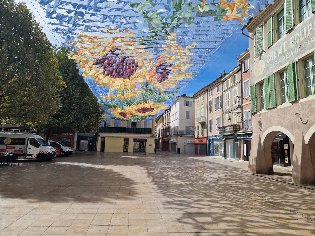 La place du Marché à Montélimar sous un voile de fanions colorés dessinant des tournesols, avec les arcades et les façades à volets verts
